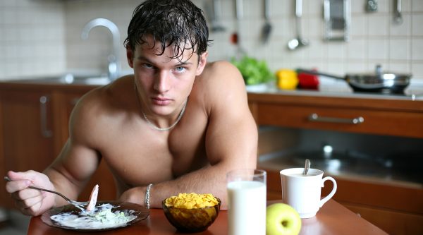 Мужчина с голым торсом и взглядом исподлобья сидит за столом с едой и держит в руке вилку